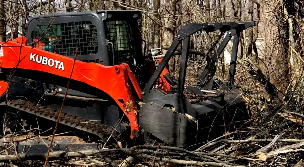 Orange Kubota excavator clearing debris in a wooded area with fallen trees and brush