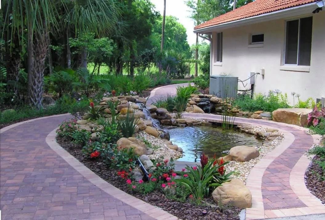 Curved stone path around a small garden pond beside a white house with red roof tiles