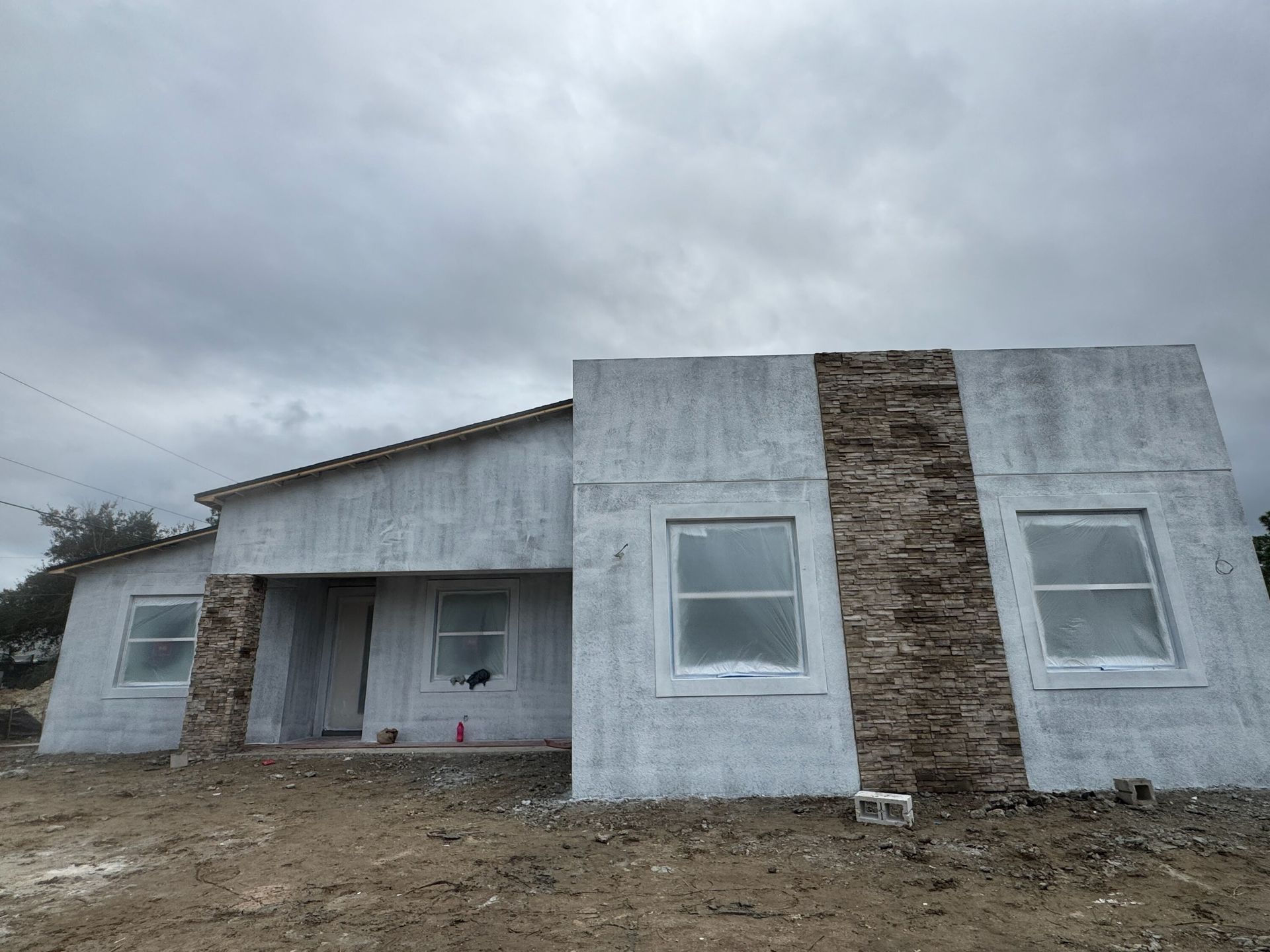 Small unfinished concrete house with front porch and stone accent under a cloudy sky