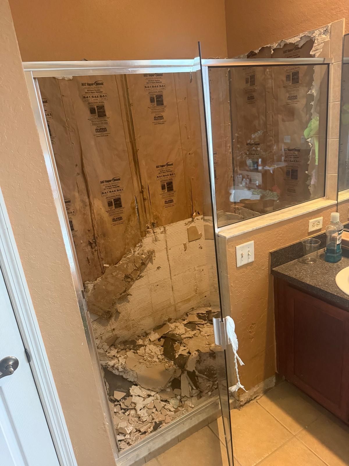 Damaged shower with missing wall panels, debris on the floor, next to a bathroom vanity and mirror.