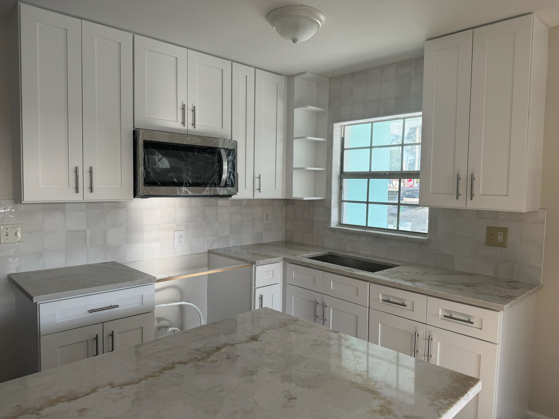 Bright white kitchen with cabinets, microwave, stove, sink, and window over a marble island.