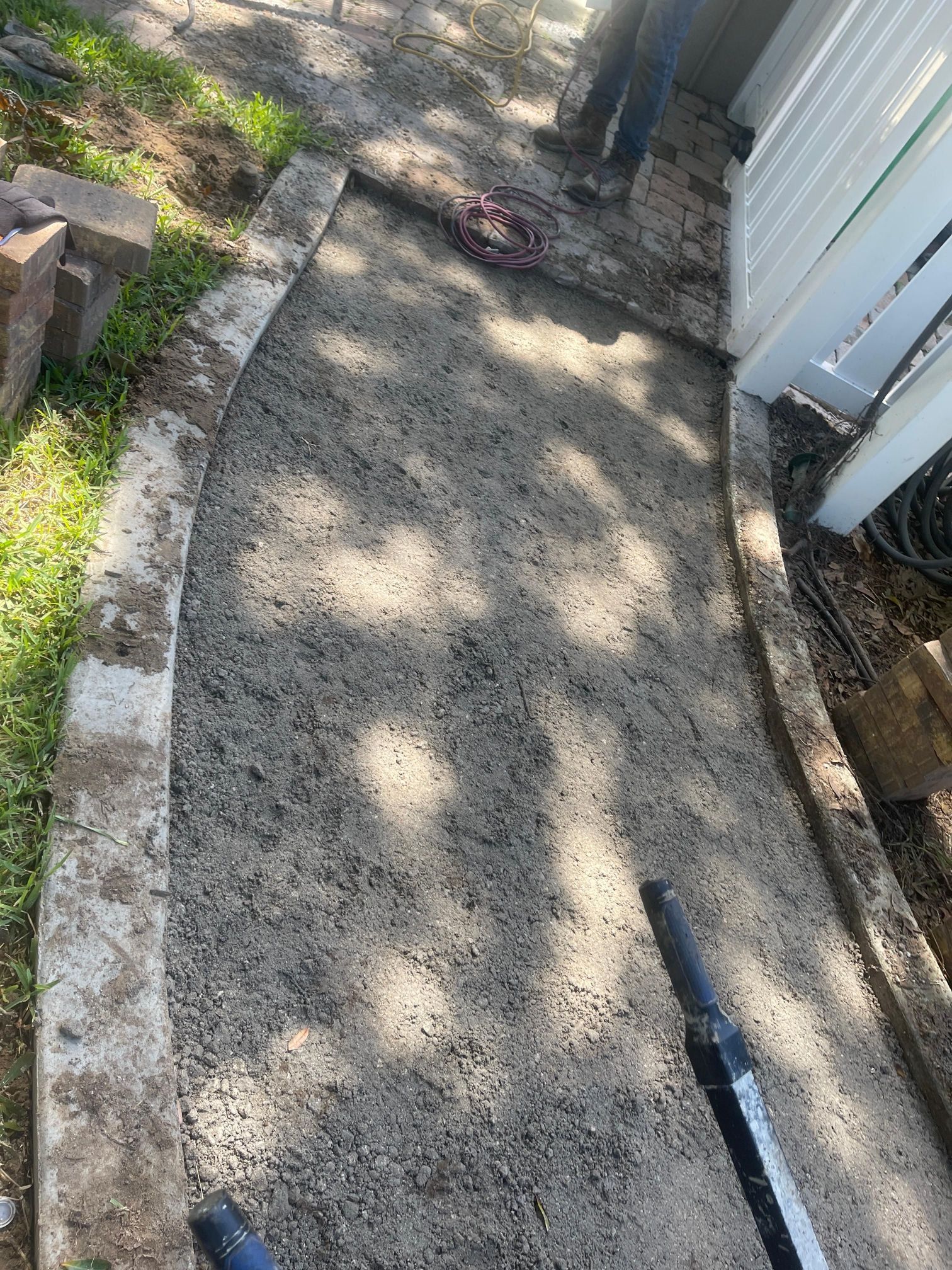 Narrow concrete walkway beside a white fence and garden bed, with scattered debris and a black hose.