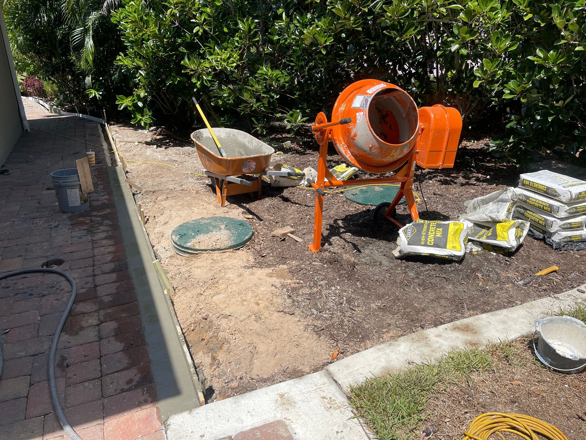 Backyard construction area with orange cement mixer, wheelbarrow, and stacked pavers on dirt ground