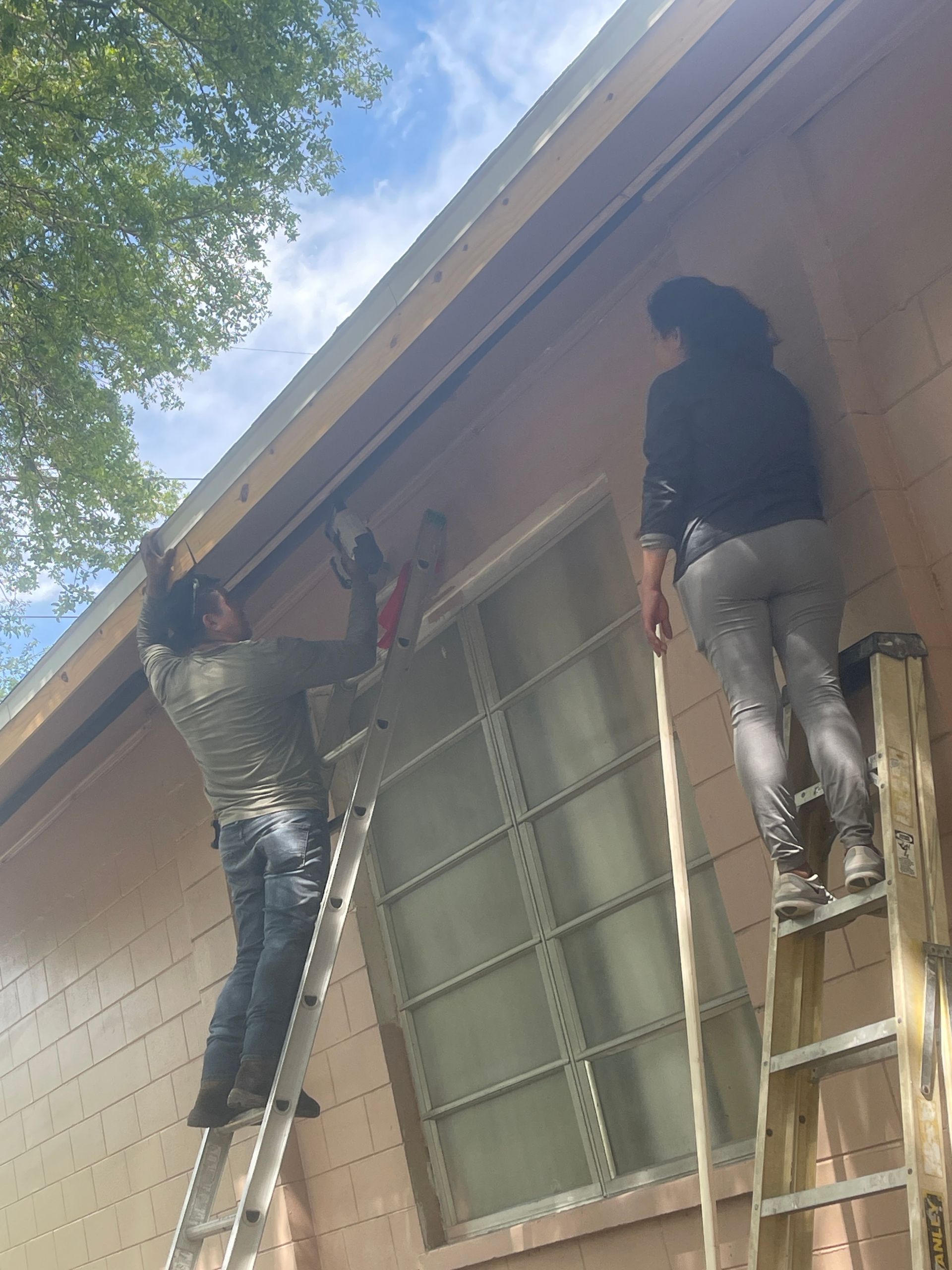 Two people on ladders working on a house exterior near a window and roofline