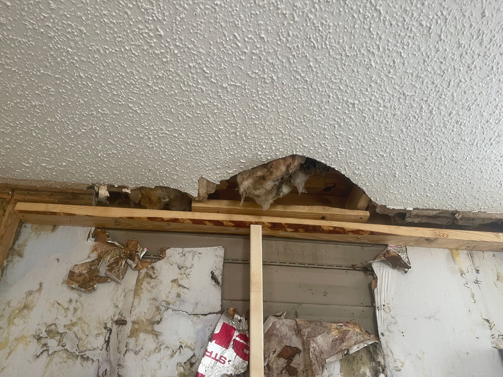 Water-damaged ceiling with exposed wood framing and torn wall covering around a repaired opening