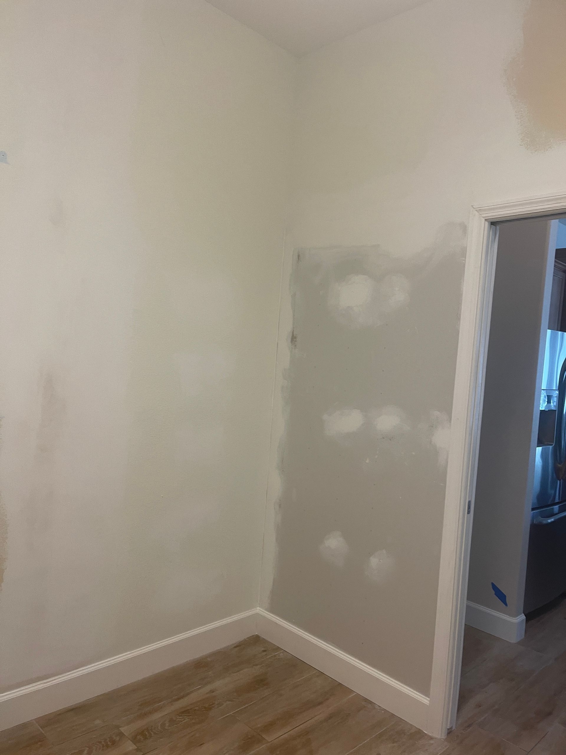 Small empty room with beige walls, patchy drywall repair, white trim, and wood flooring near a doorway.