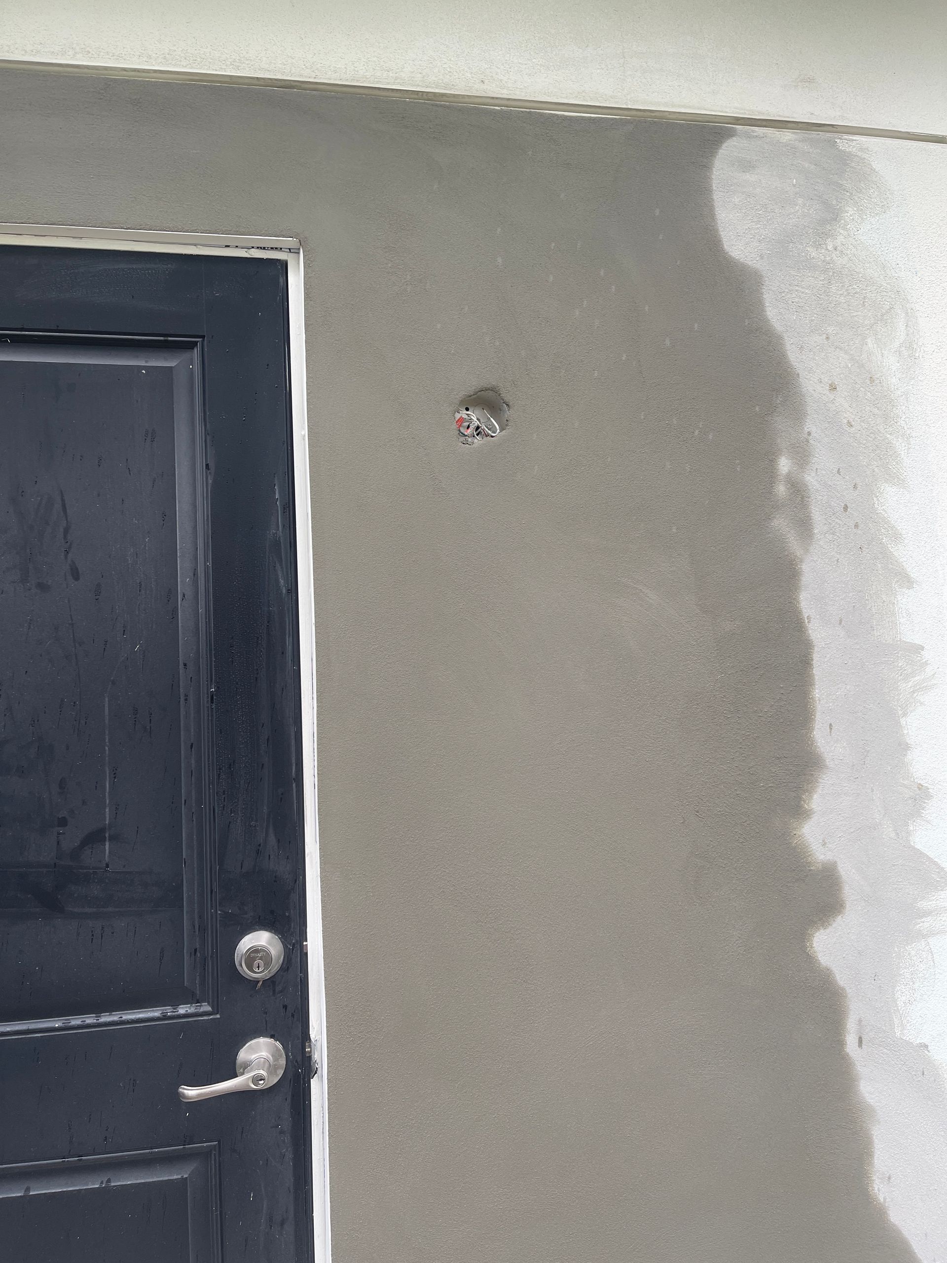 Gray wall beside a black door with a small hole near the center and rough white plaster on the right edge