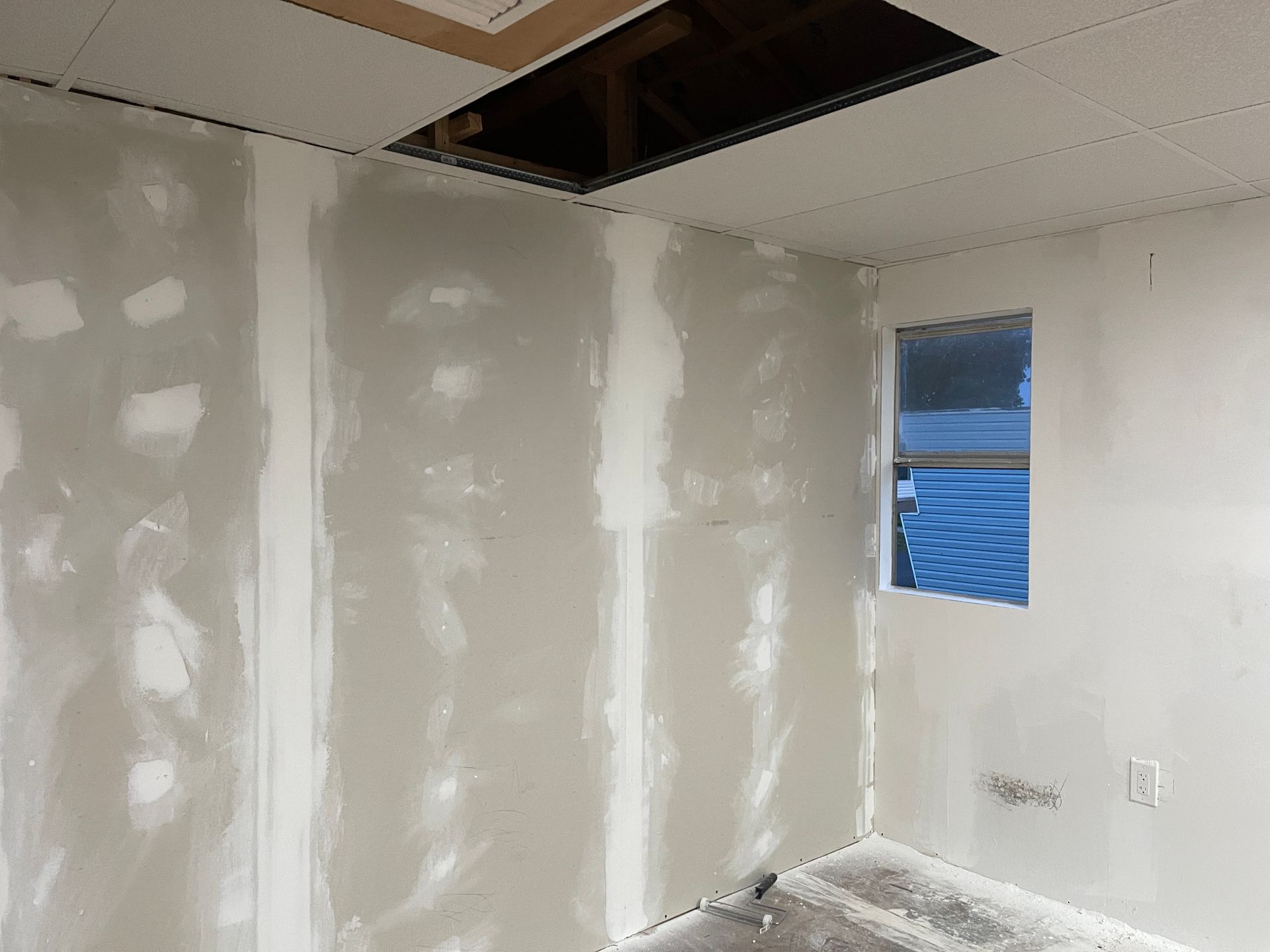 Partially finished white room with drywall patches, open ceiling panel, and a small window.