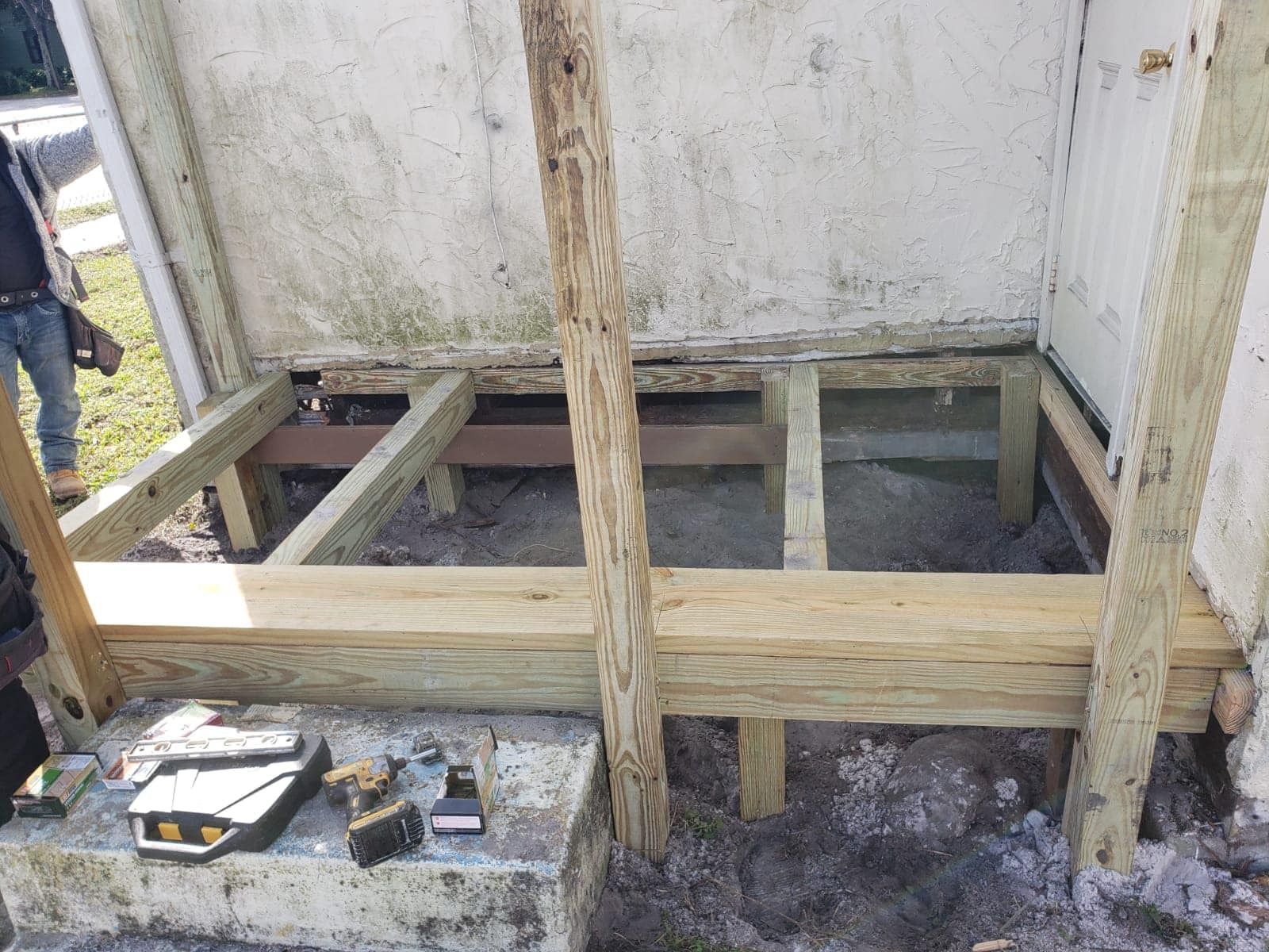 Partially built wooden deck frame beside a white wall, with tools on the ground and a person at left edge