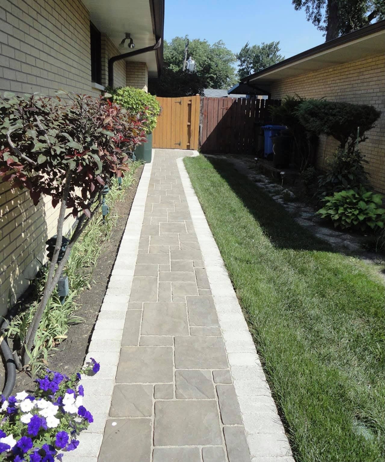 Narrow brick walkway between houses leading to a wooden gate, bordered by grass and shrubs.