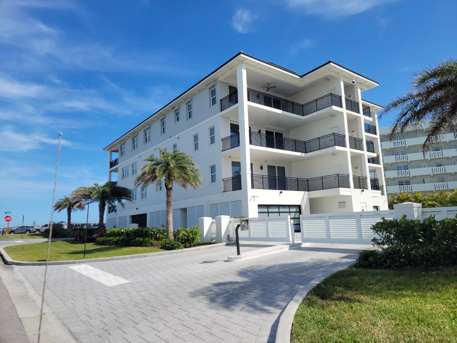 Modern white beachfront condo with balconies, palm trees, and a paved driveway under a blue sky