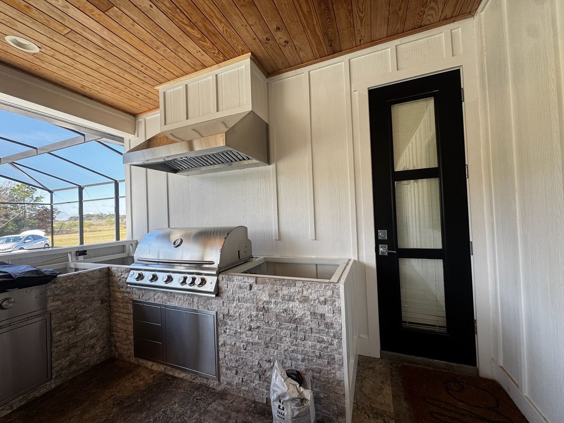 Covered outdoor kitchen with stainless grill, stone counters, wood ceiling, and black door.