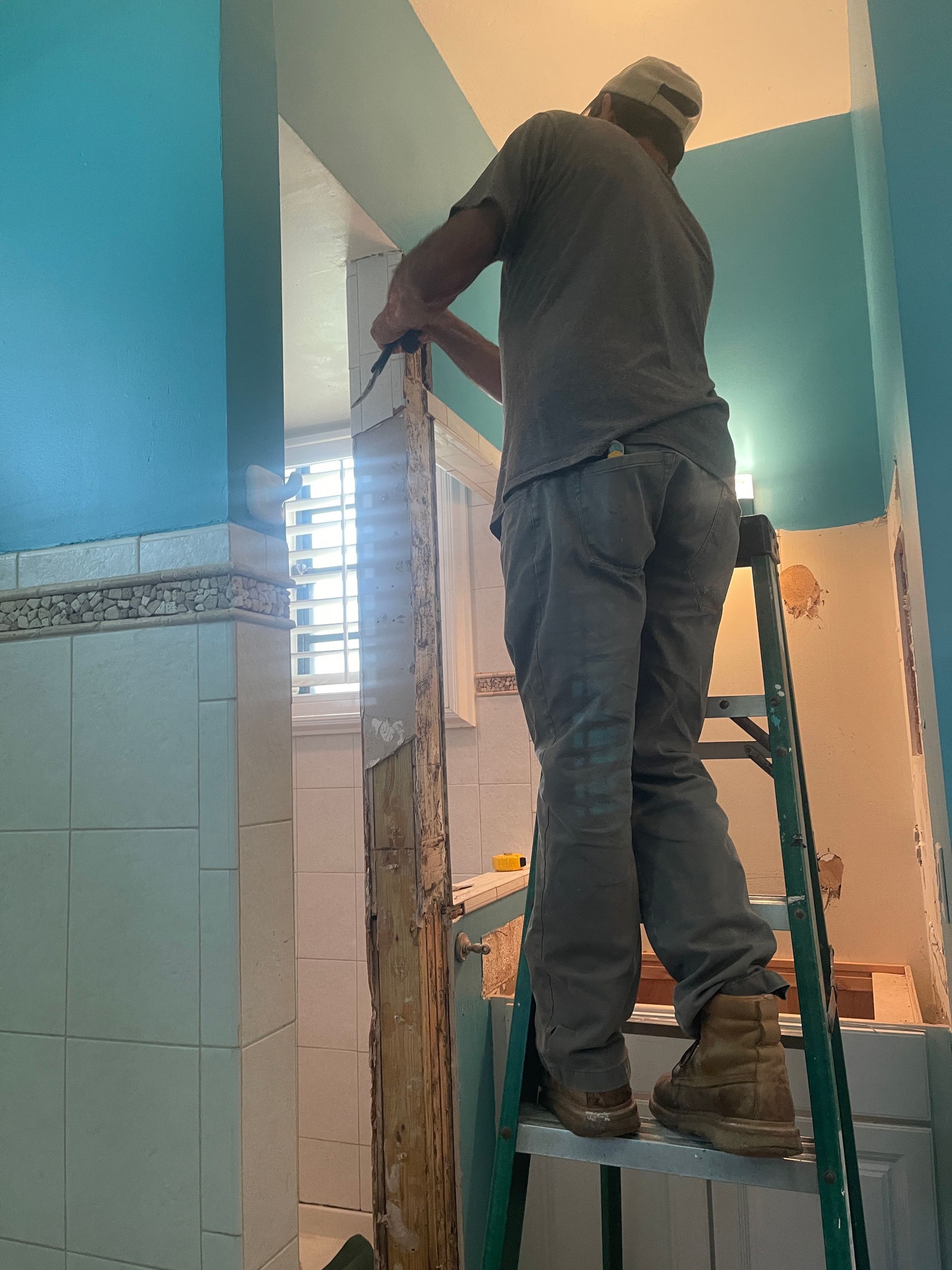 Worker standing on a ladder repairing a wall in a teal-and-tile bathroom doorway