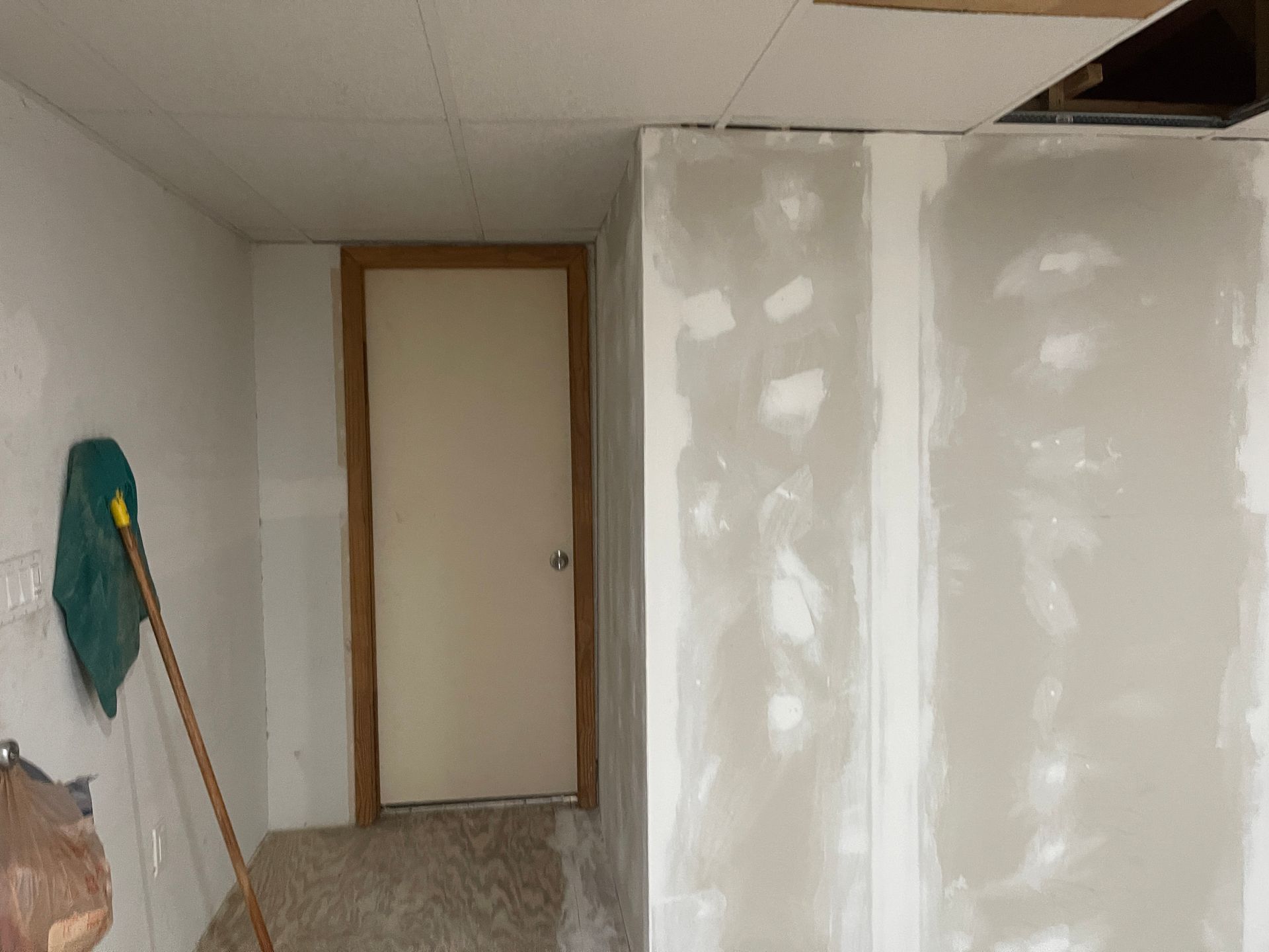 Small room under renovation with white drywall, a wooden door, and a vacuum leaning against the wall.