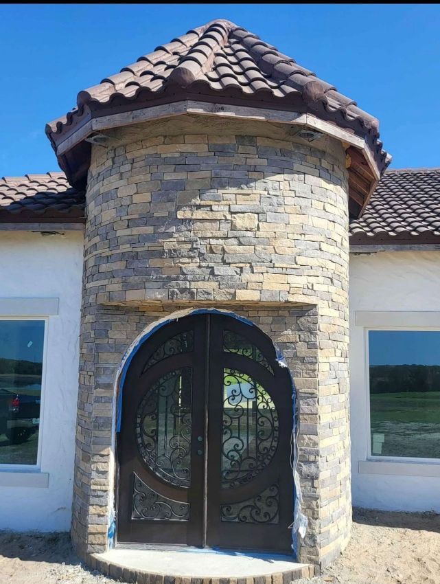 Stone turret entrance with arched black wrought-iron door and tiled roof on a stucco house
