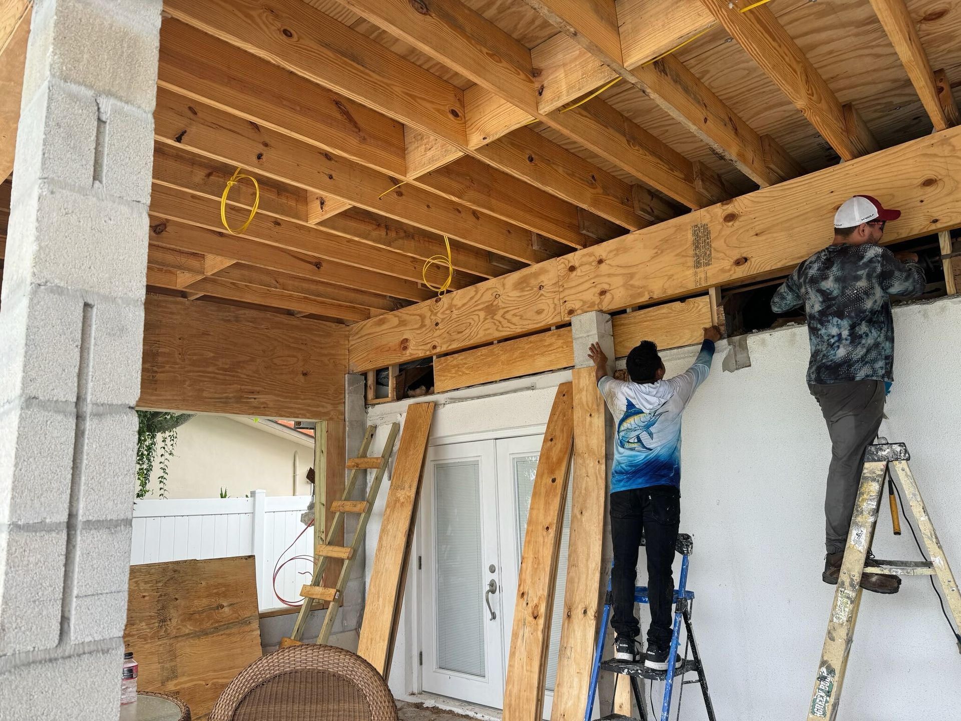 Workers installing beams under a wooden porch ceiling, with ladders and a white double door below.