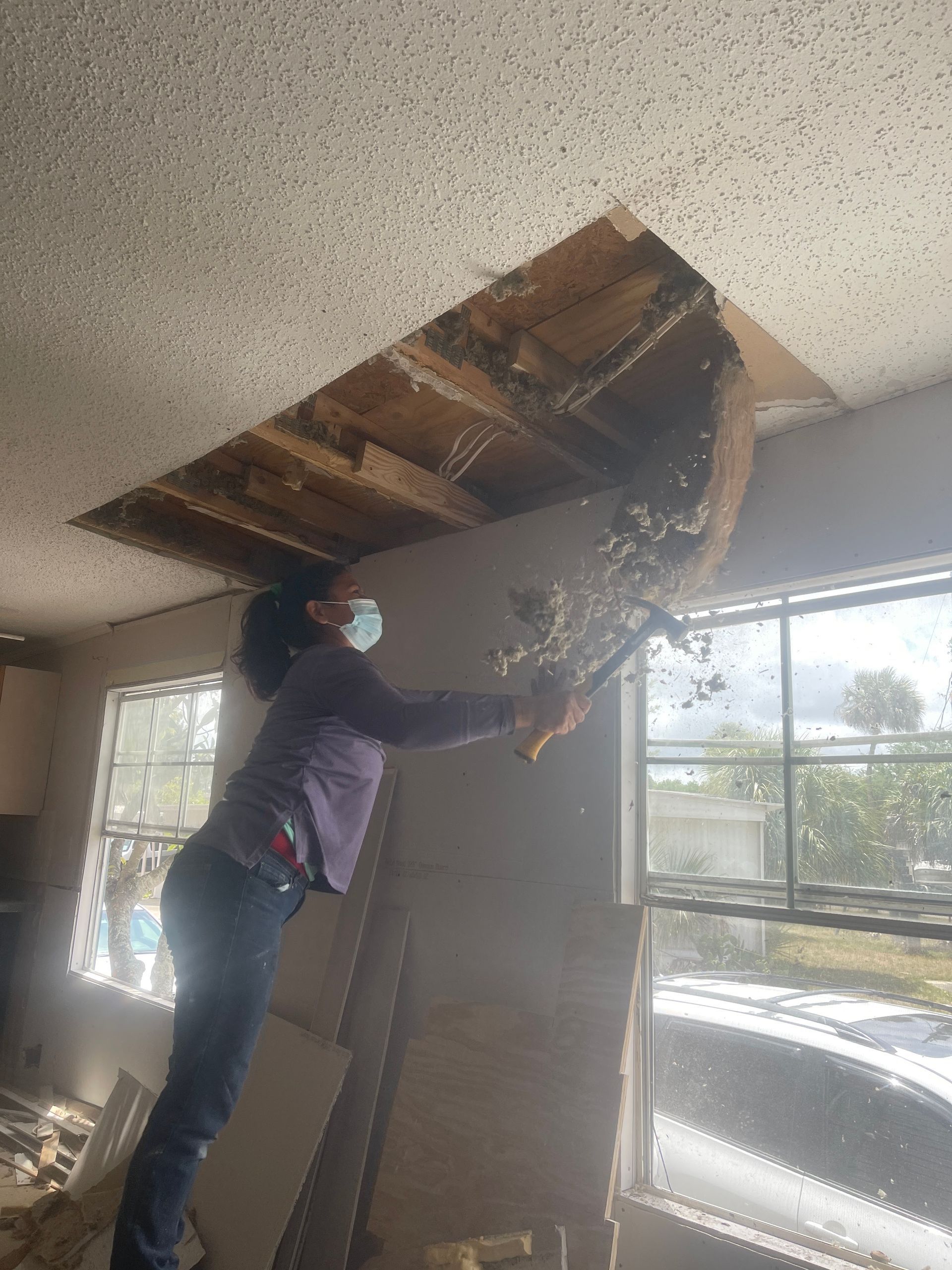 Person removing damaged ceiling panel with a ladder in a room with large windows.