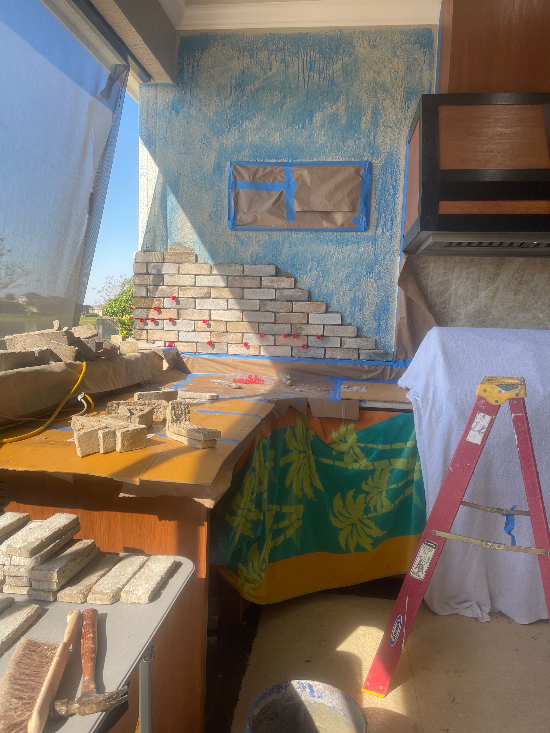 Sunlit room with blue textured wall, ladder, table clutter, and exposed brickwork under repair
