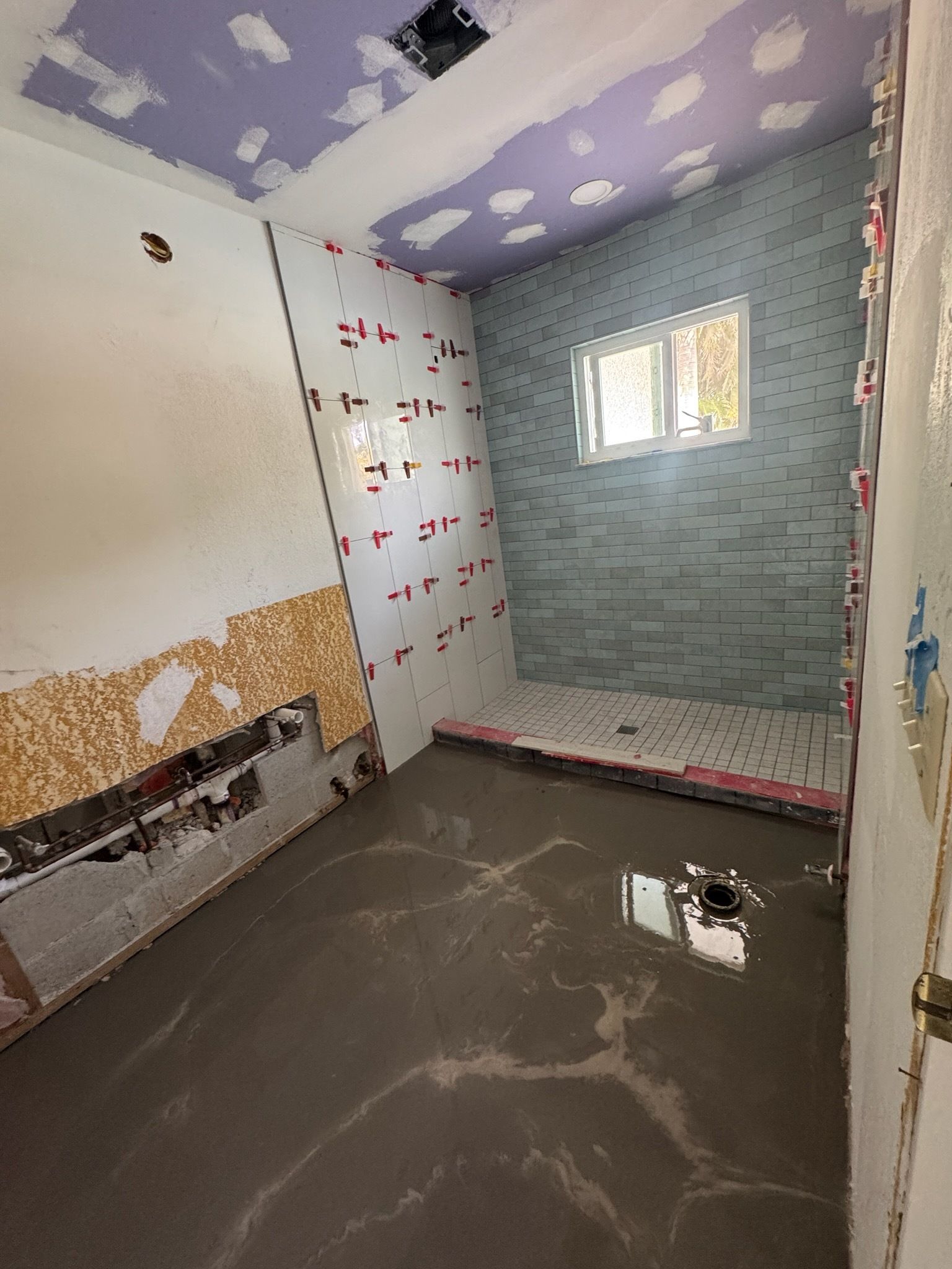Small unfinished bathroom with tiled walls, purple ceiling, window, and exposed floor drain.