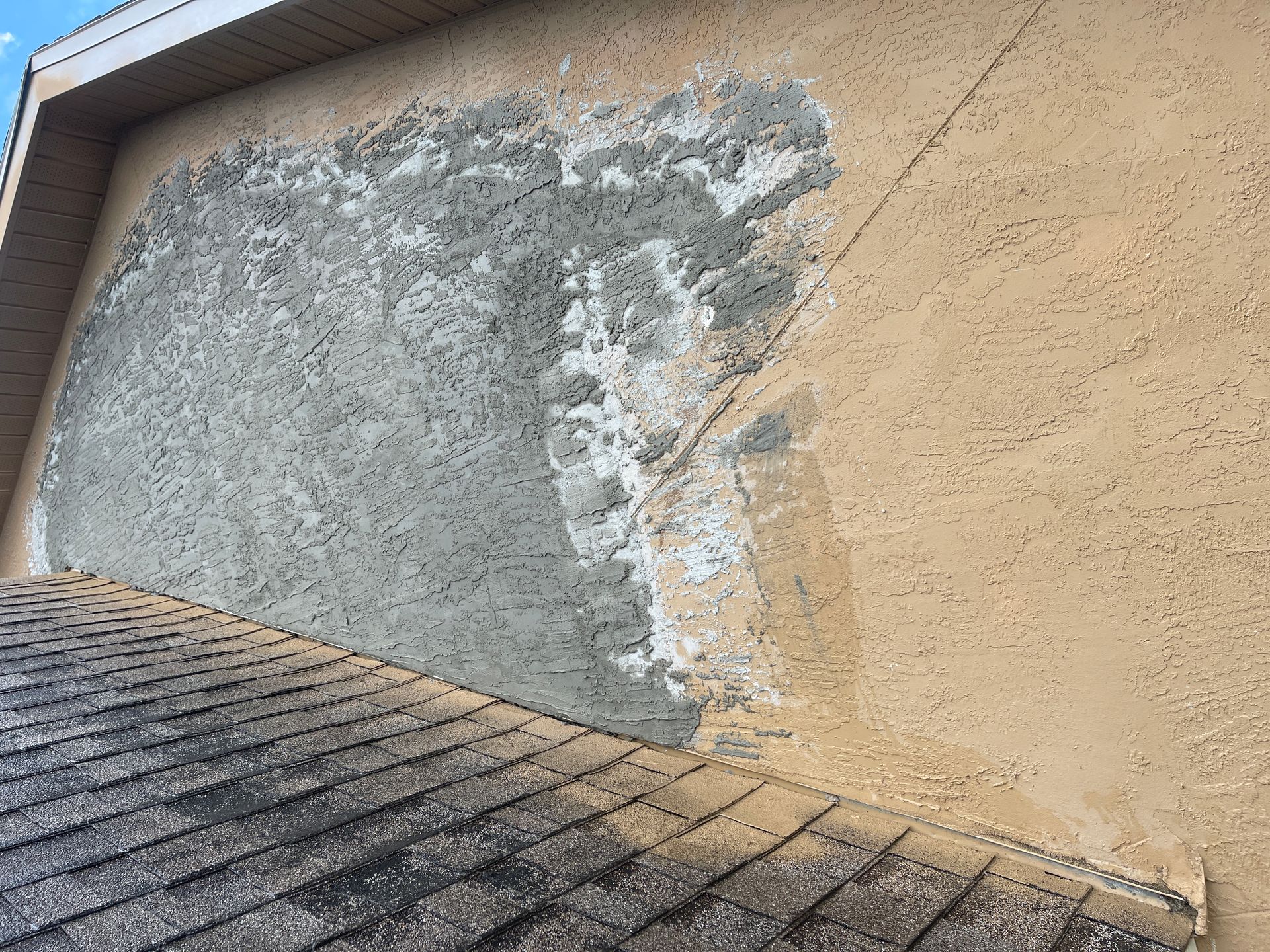 Damaged exterior stucco wall beside a shingled roof, with a large gray patch and exposed repair area
