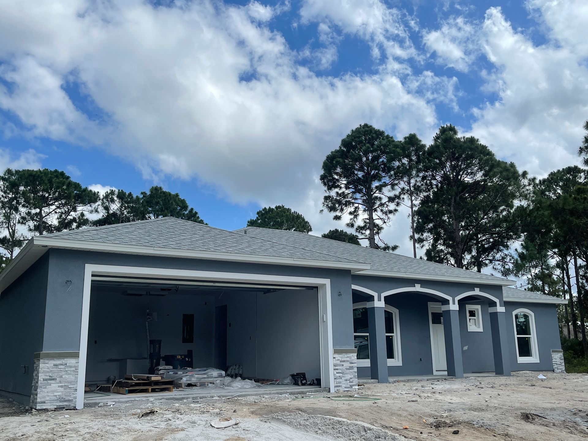 Gray single-story house under construction with open garage, white trim, and cloudy sky