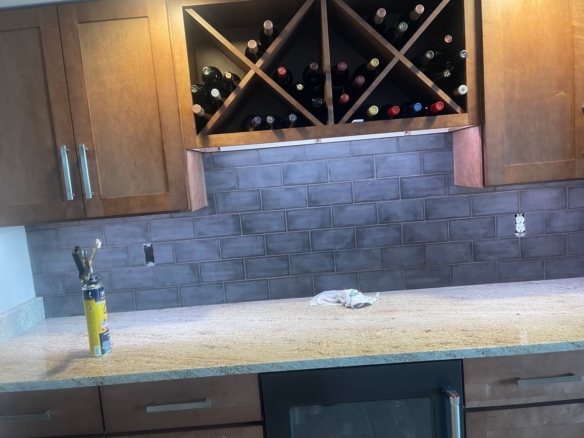 Kitchen with dark cabinets, wine rack, gray backsplash, speckled countertop, and cleaning supplies on the counter