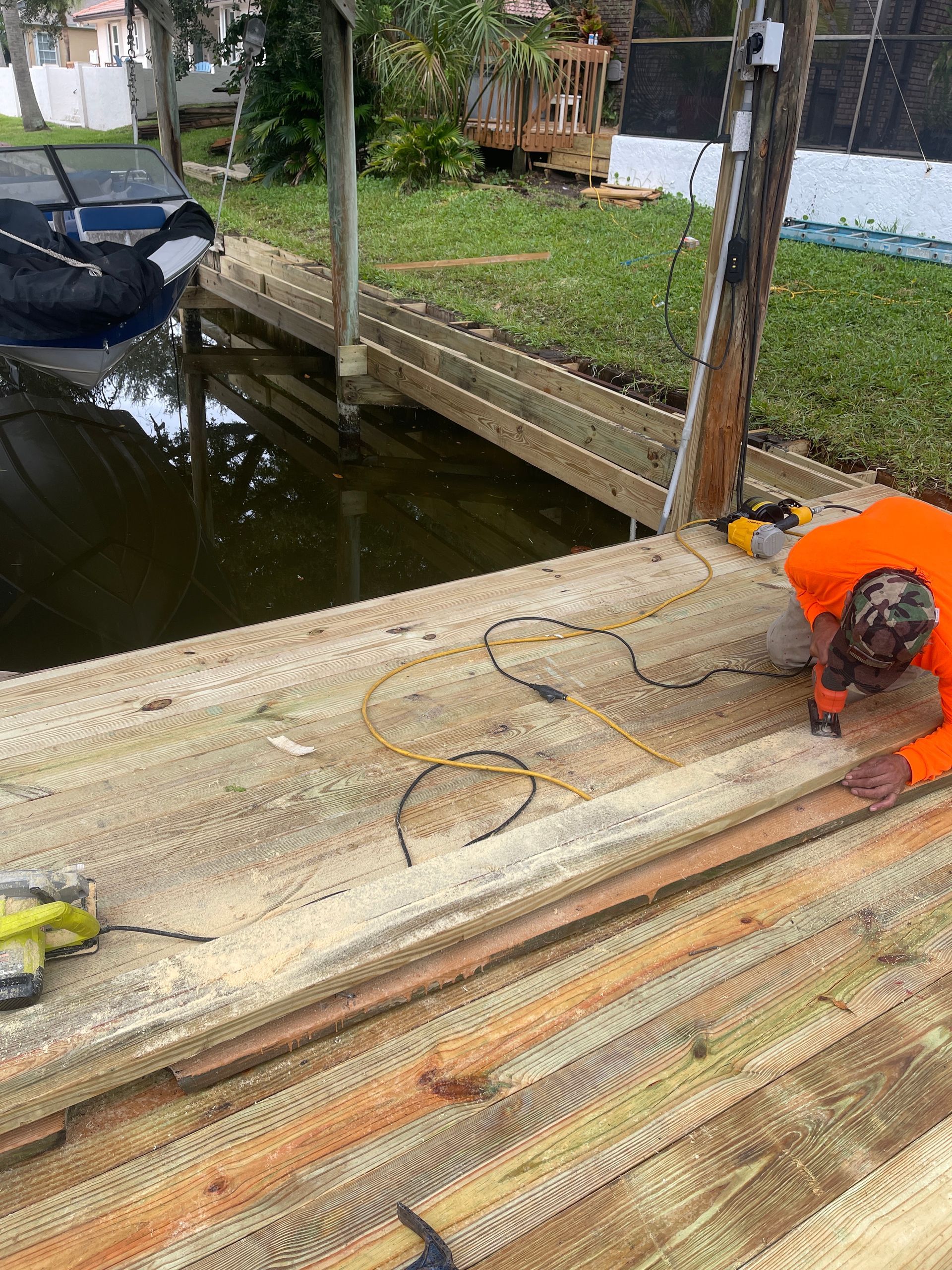Worker in orange shirt crouches on wooden dock by a canal, cutting or sanding boards near a parked boat.