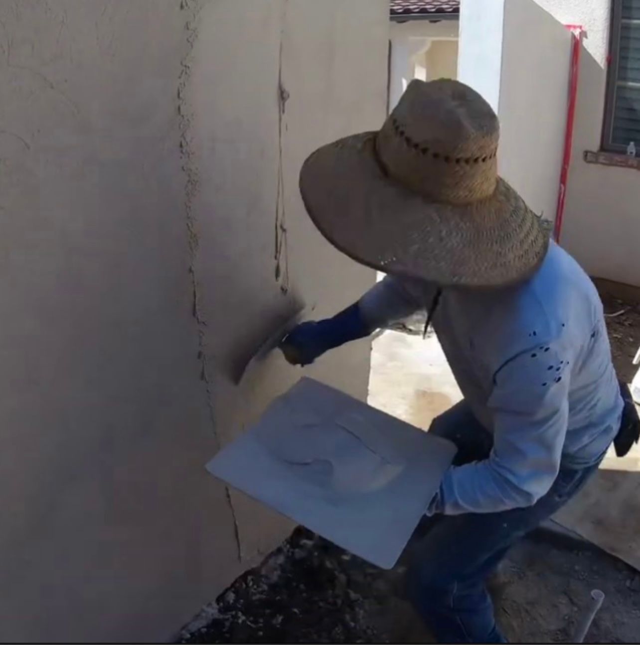 Person in a straw hat applying paint to a wall while holding a tray of plaster or paint