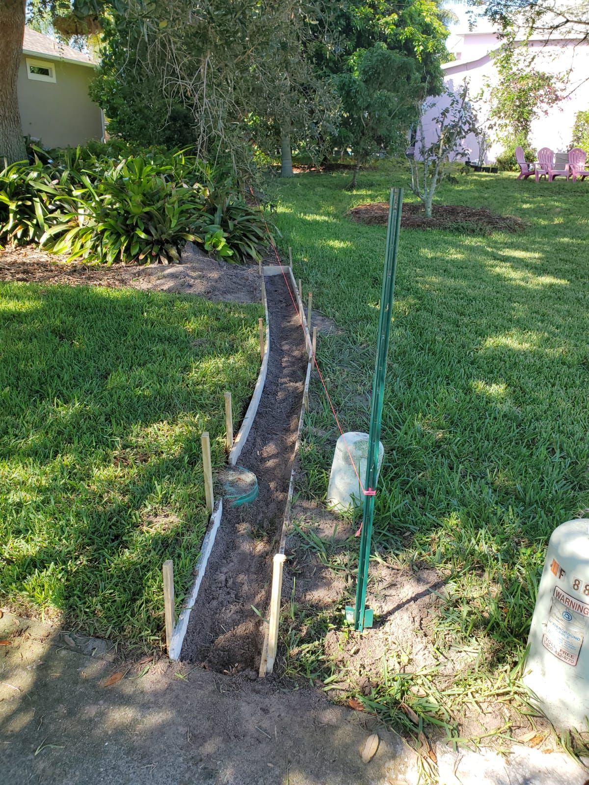 Garden path under construction beside a grassy yard with a trench, stakes, and pipes