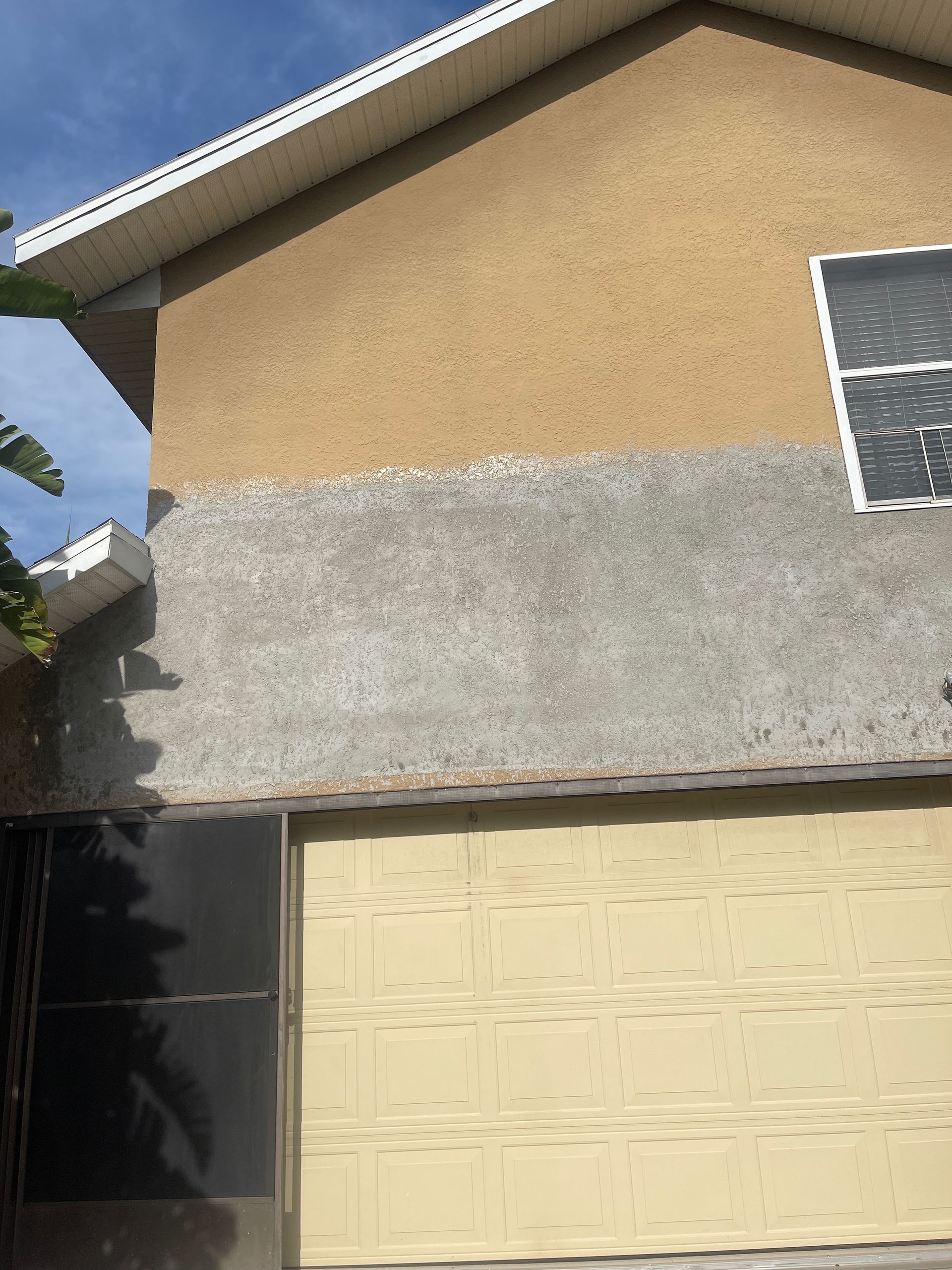 Two-story house exterior with tan stucco, gray lower wall, garage door, and a small window.