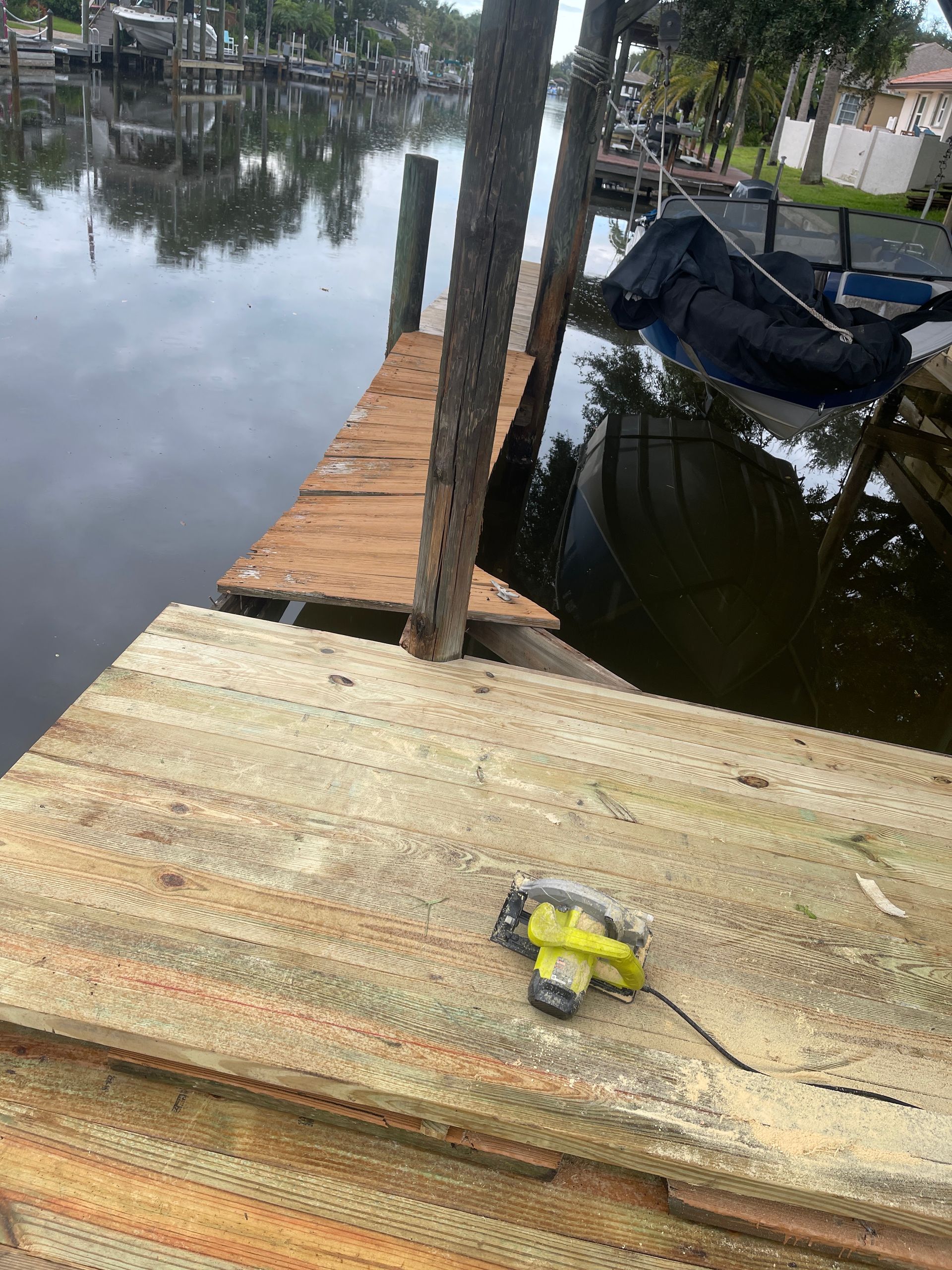 Wooden dock by a calm canal, with a yellow tool on the foreground and boats to the right