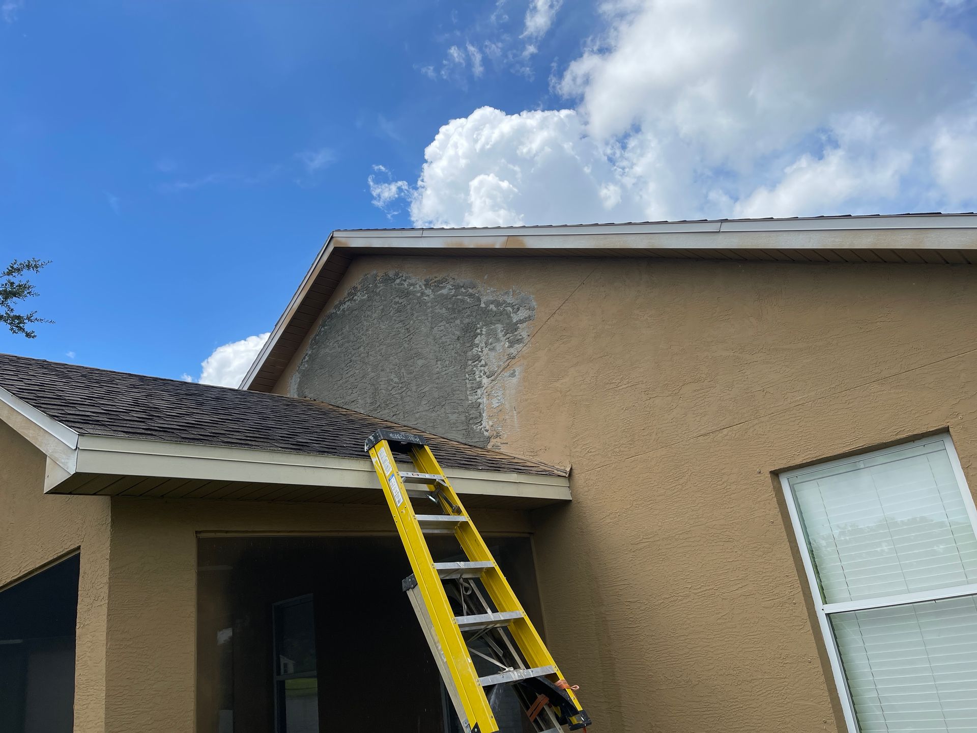 Ladder leaning against a tan house under roof damage with missing stucco near the eave