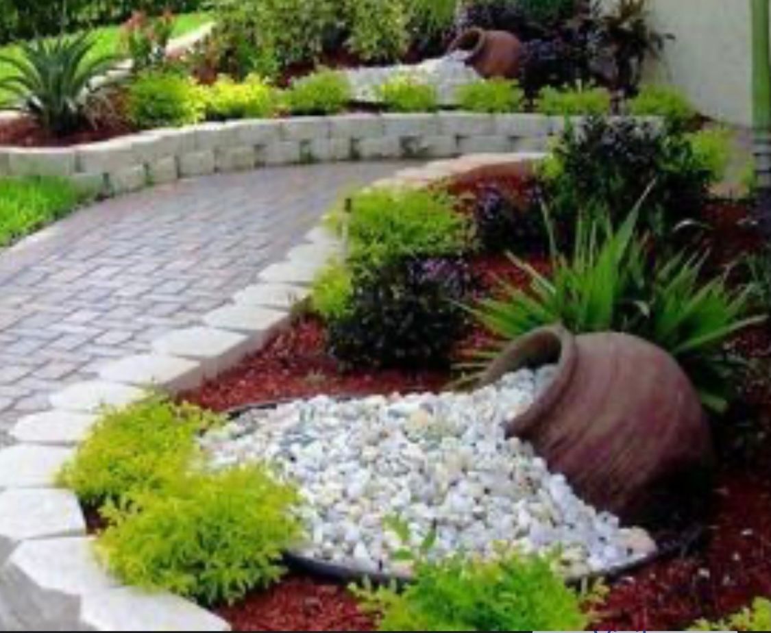 Curved stone path through a landscaped garden with green plants, red mulch, and white rocks by a terracotta pot