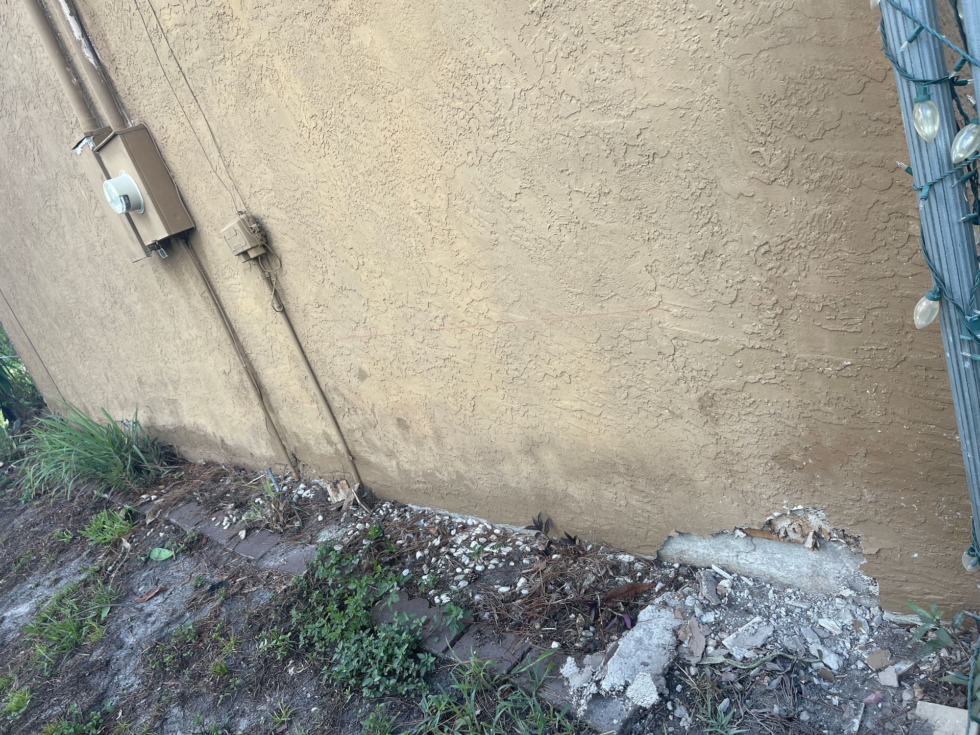 Dirt and weeds along a beige wall beside a chain-link fence, with a damaged concrete patch.