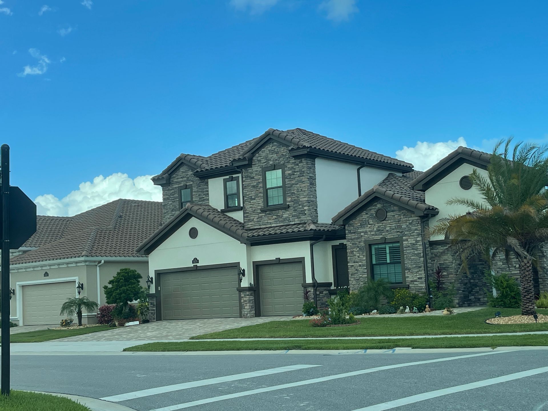 Suburban two-story stone house with a tiled roof, garage, and neatly kept lawn under a blue sky