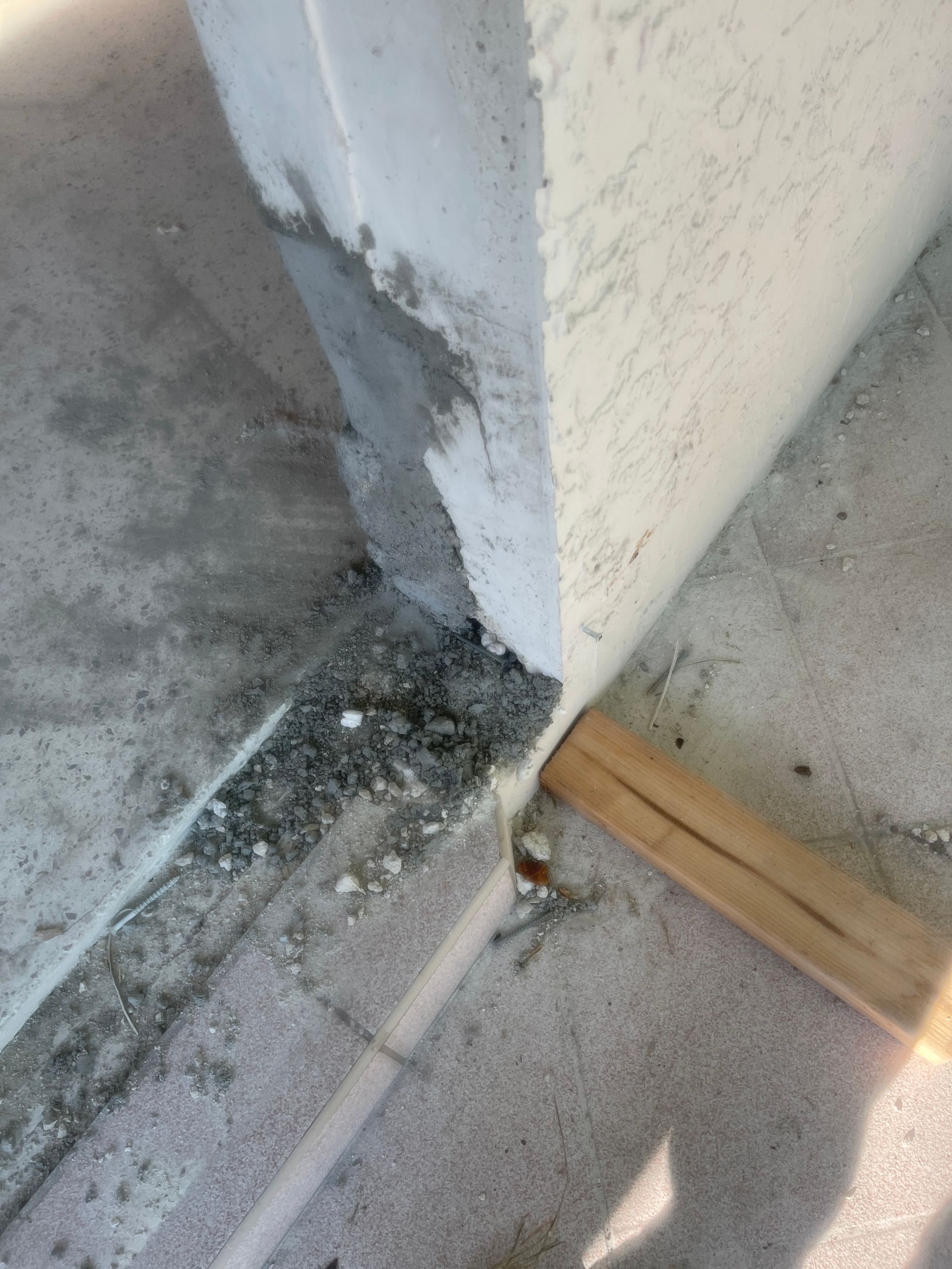 Damaged concrete wall corner with chipped plaster and debris on a gray floor near a wood strip