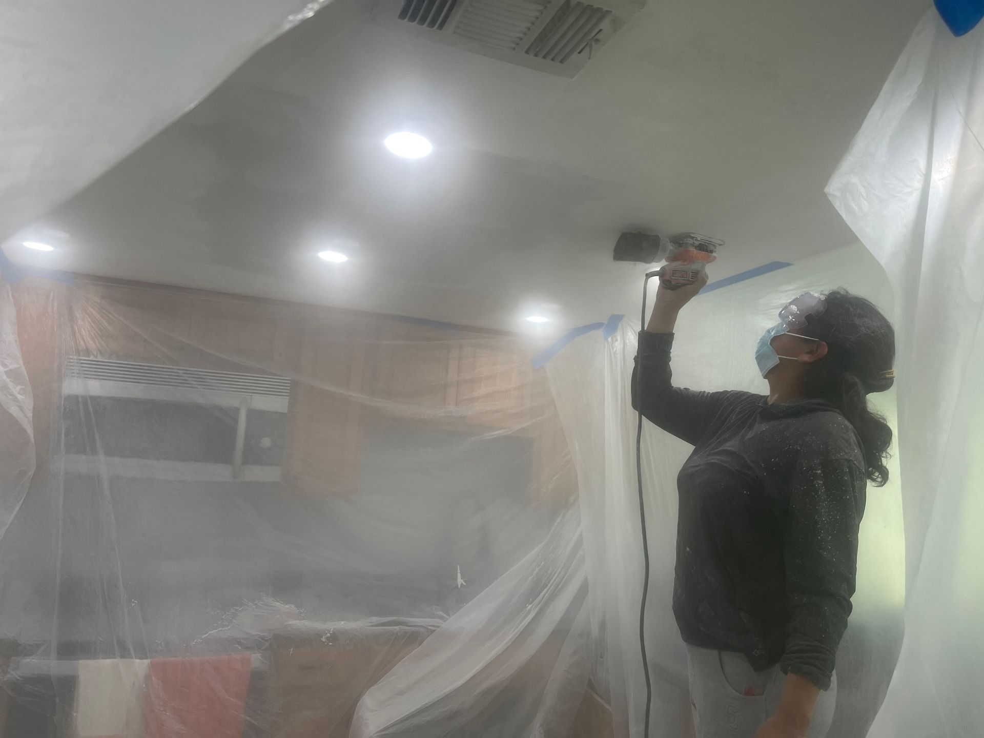 Person using a drill  while standing on a ladder in a room under renovation, with plastic sheeting and ceiling lights.