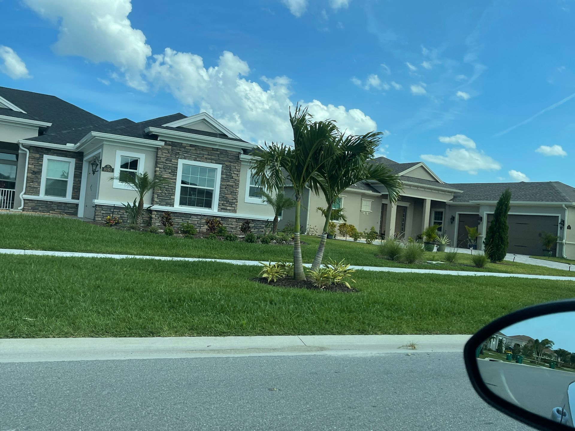 Modern suburban house with manicured lawn and palm tree under a blue sky, viewed from a car.