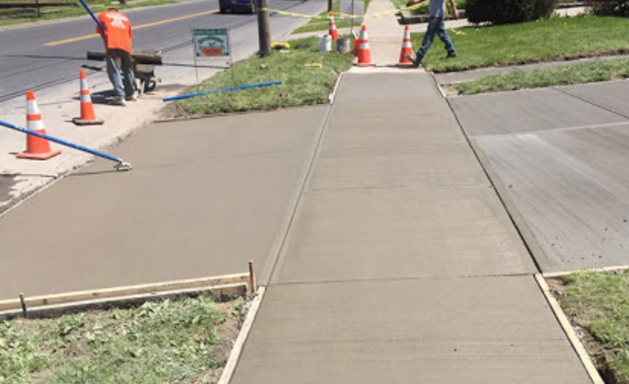 Concrete sidewalk under construction with orange cones and a worker near the road