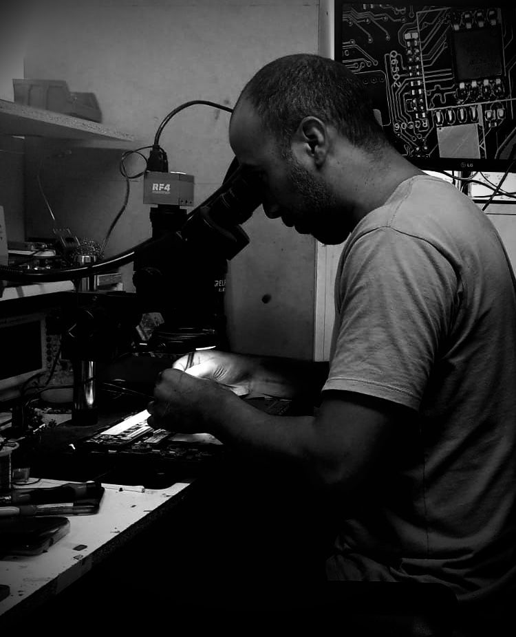 Hombre que utiliza un microscopio, enfocado en una placa de circuito, en un taller poco iluminado.