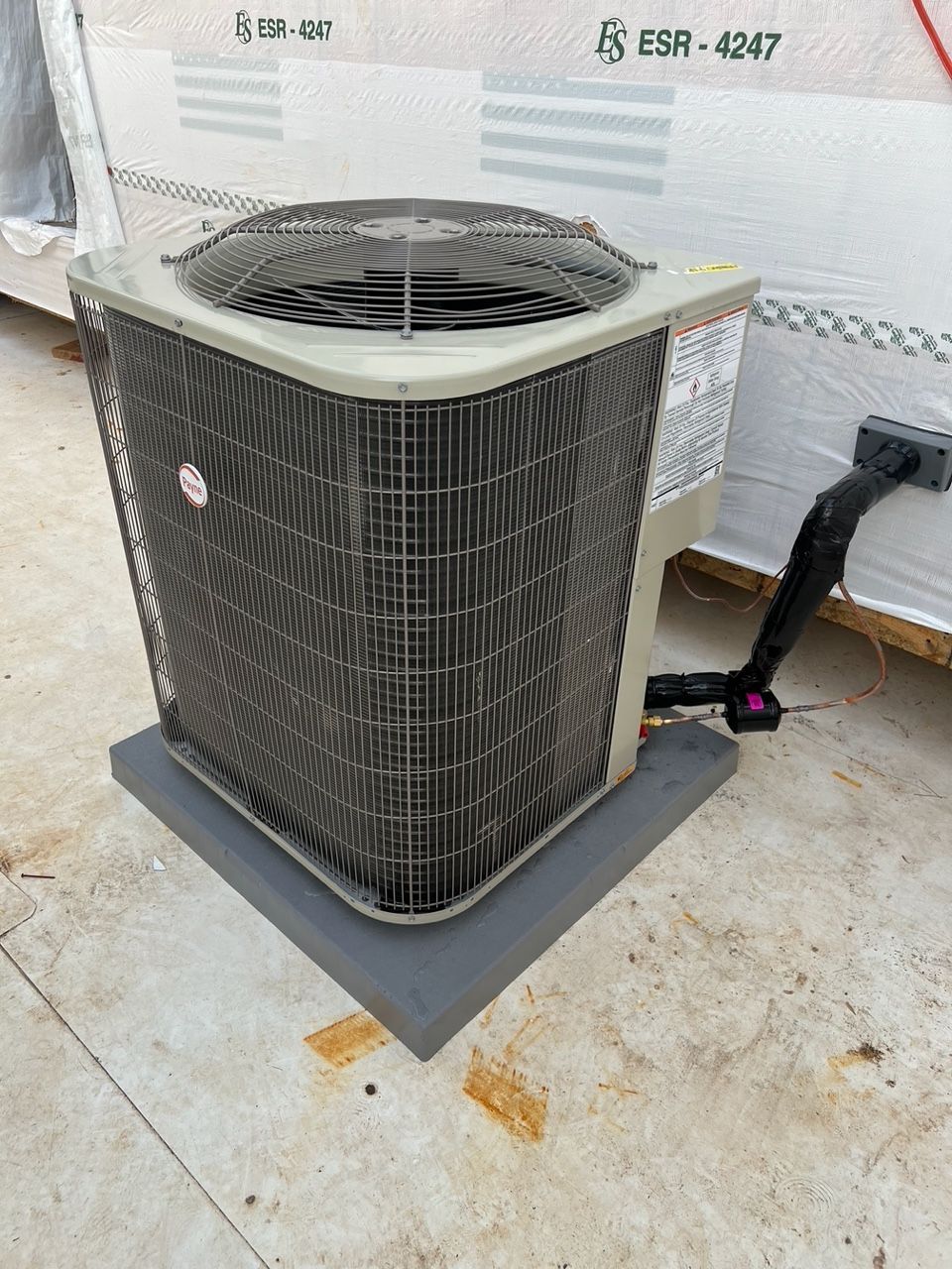 An outdoor residential air conditioning unit sitting on a grey plastic pad against a building wall.