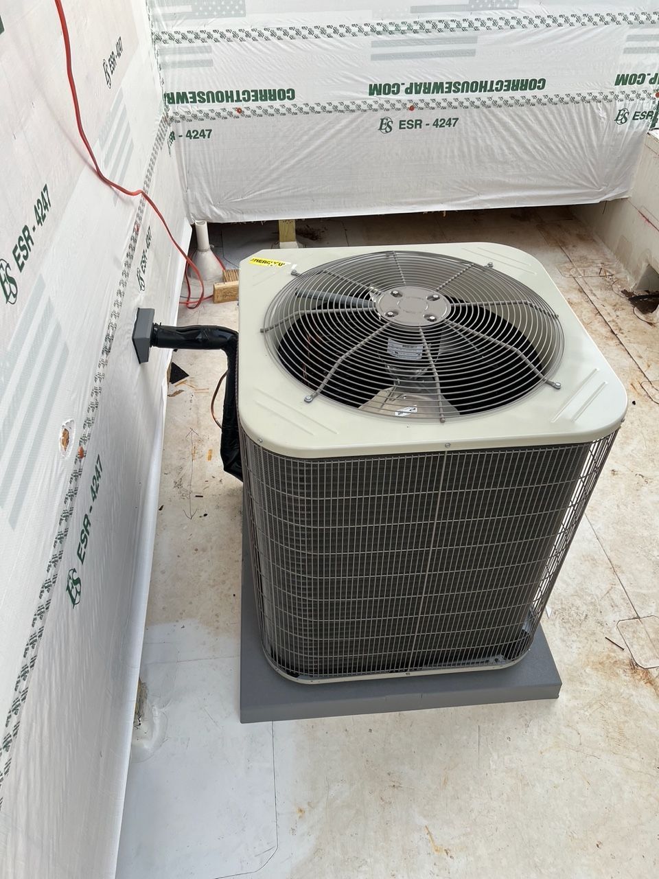 An outdoor AC condenser unit sits on a gray pad next to a building wall covered in white construction wrap.