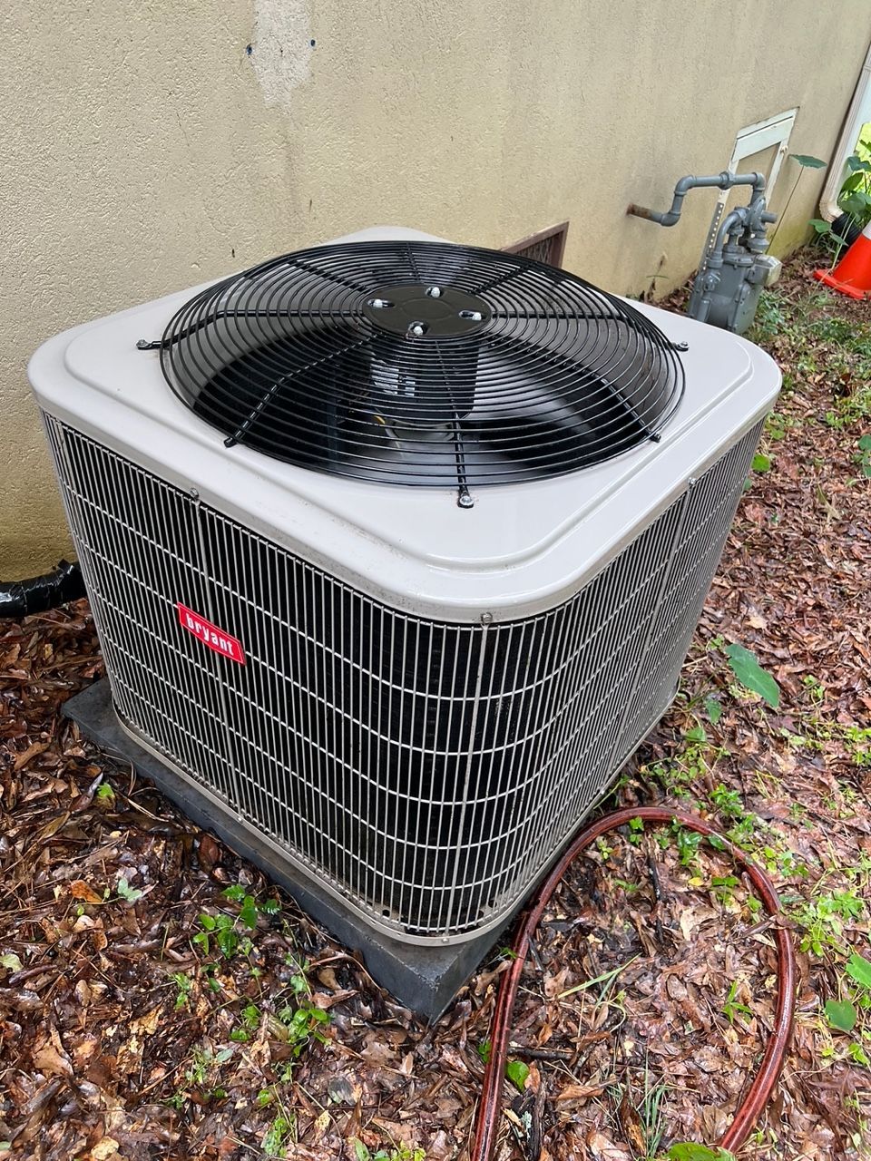 A square, grey outdoor HVAC condenser unit sits on a concrete pad surrounded by mulch and leaves next to a tan wall.