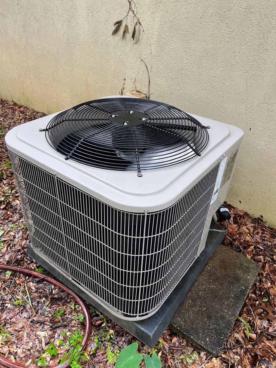 A grey outdoor air conditioning unit sits on a concrete pad against a beige wall, surrounded by dry leaves and greenery.