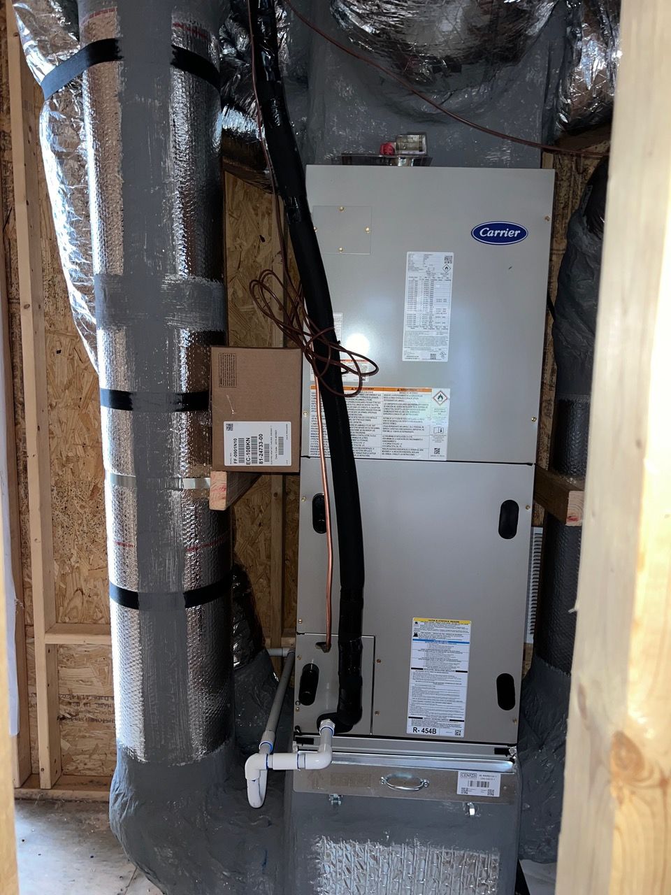 A grey Carrier HVAC unit installed in an unfinished wooden closet with silver insulated ductwork on the sides.
