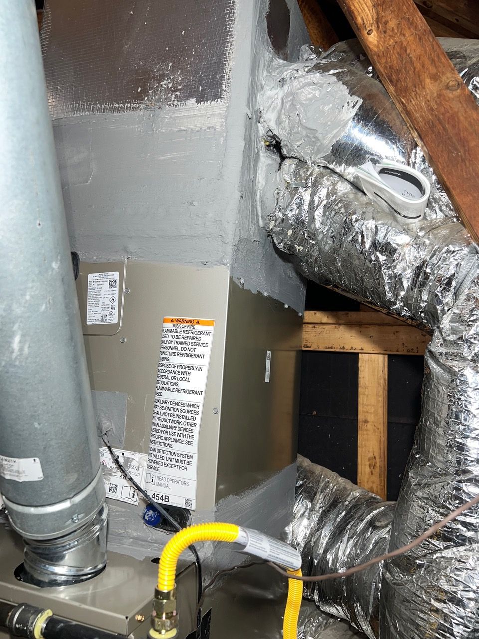 An HVAC furnace unit with a metal exhaust pipe and foil-covered flexible ductwork in an attic setting.