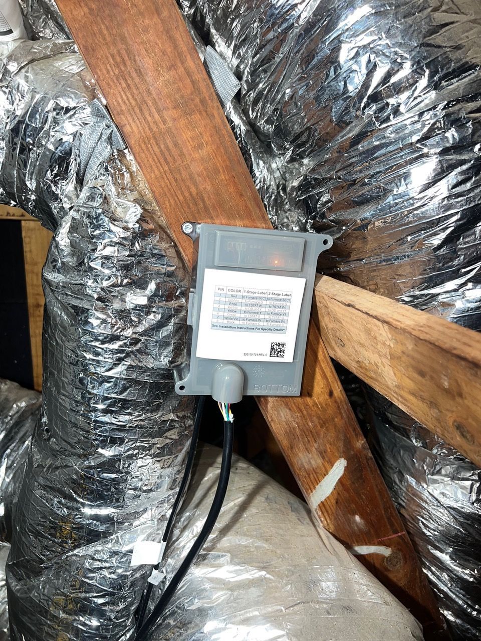 A gray electronic box is mounted to a wooden rafter in an attic, surrounded by silver-insulated HVAC ducts.