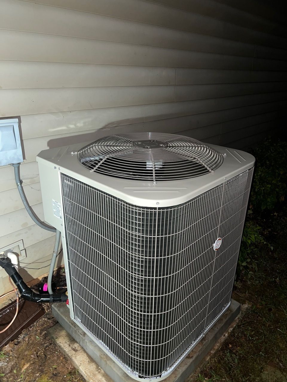 A light-colored outdoor air conditioning unit sits on a concrete pad next to a house with beige siding.