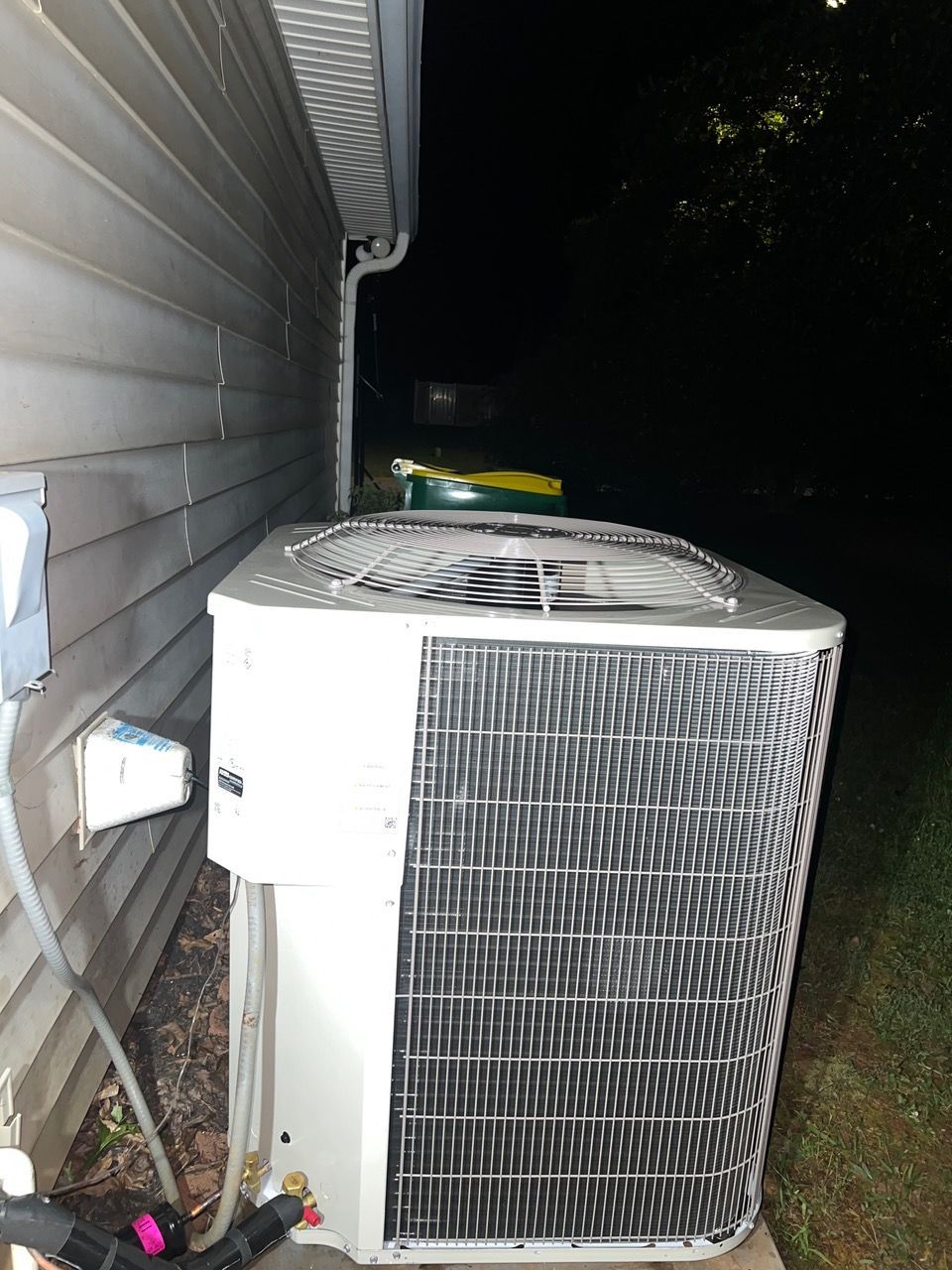 An outdoor air conditioning unit sits next to a light-colored vinyl-sided house at night.