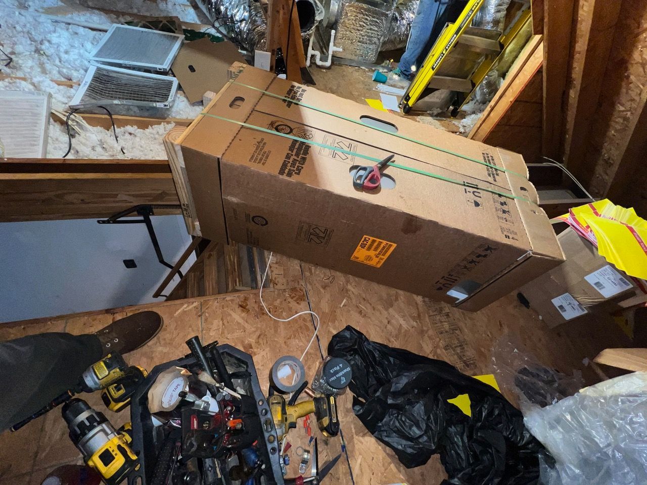 A box and tools in an attic space with wood flooring, open insulation, and a yellow ladder in the background.